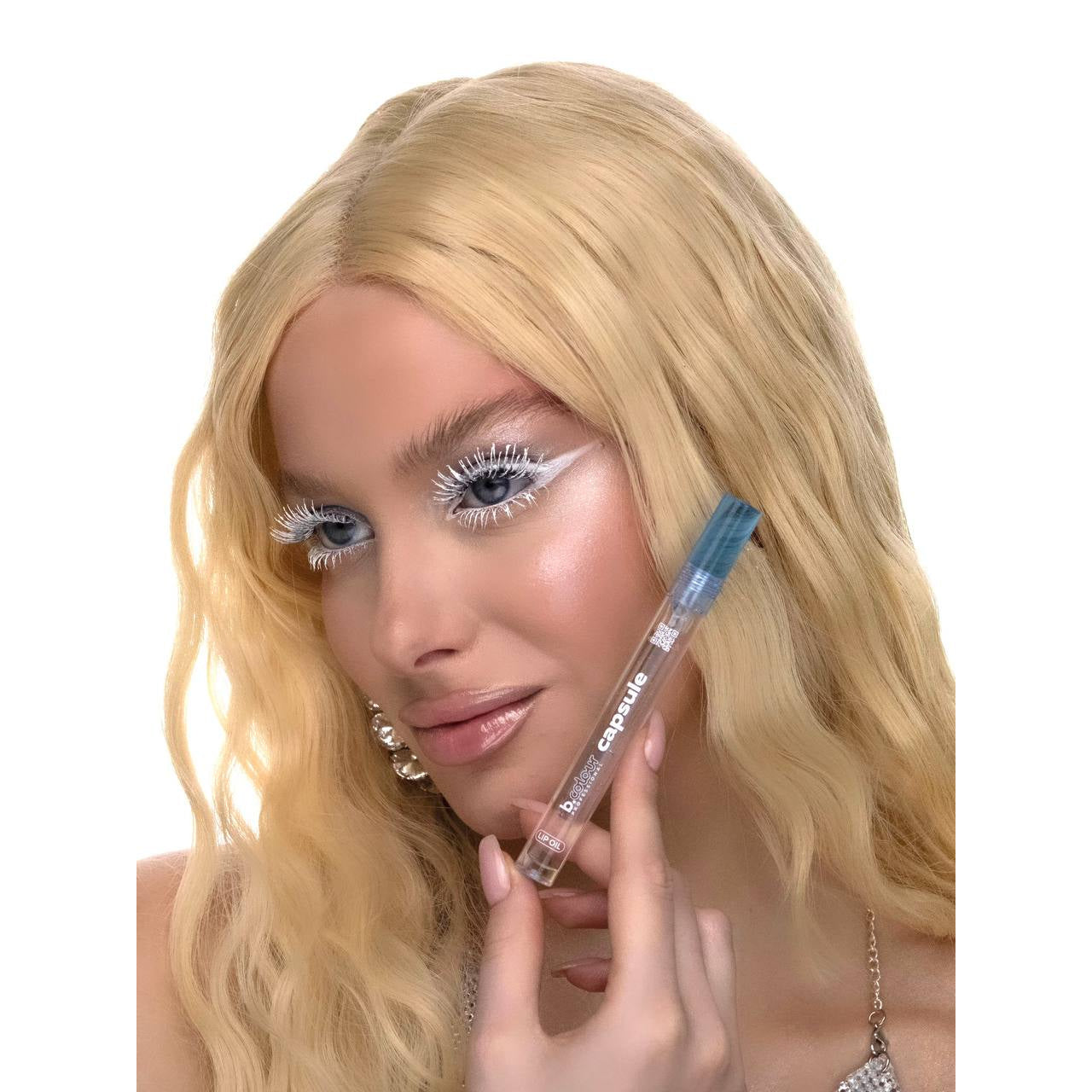 Frau mit langem blondem Haar und auffälligem weißem Augen-Make-up hält das schlanke Lipgloss-Fläschchen b.colour professional Lip Oil Capsule mit blauem Deckel in der Hand, ihre Lippen glänzen natürlich.