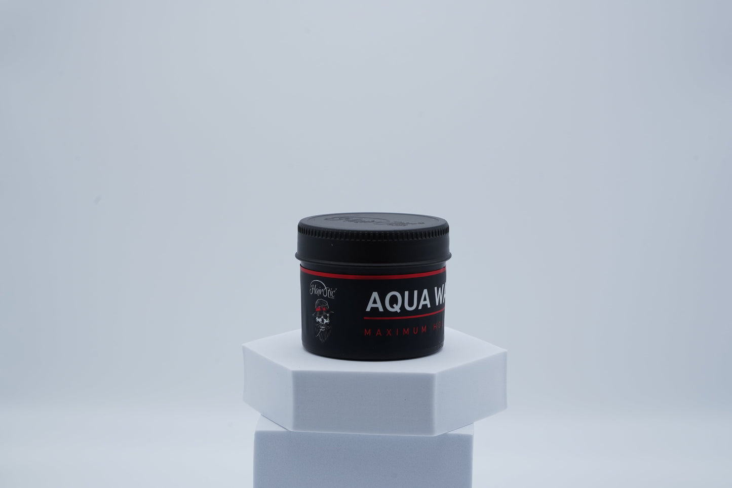 Schwarze Dose Hairotic Aqua Wax Maximum Hold mit rotem Akzent, seitlich auf weißem Podest vor neutralem Hintergrund fotografiert.
