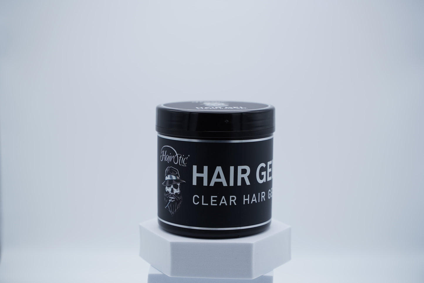 Schwarze Dose Hairotic Clear Hair Gel mit weißem Schriftzug und Logo, auf weißem Podest vor neutralem Hintergrund platziert.