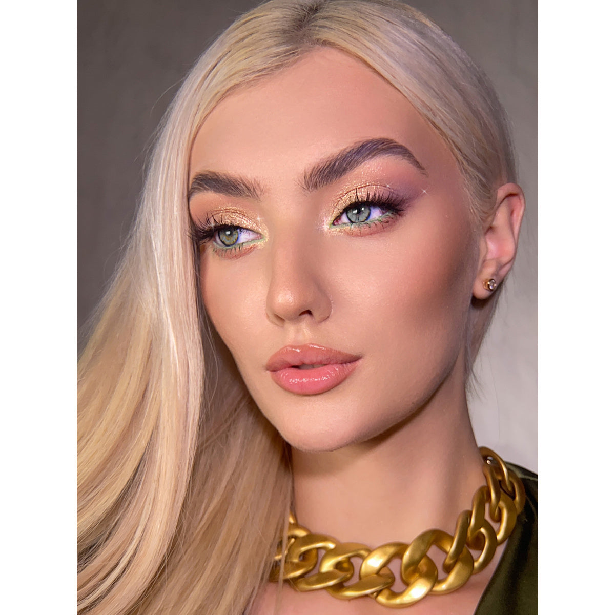 Frau mit langen blonden Haaren trägt ein glamouröses Make-up mit goldenem Schimmer-Lidschatten und dezentem Nude-Lippenstift, kombiniert mit einer auffälligen goldenen Gliederkette.