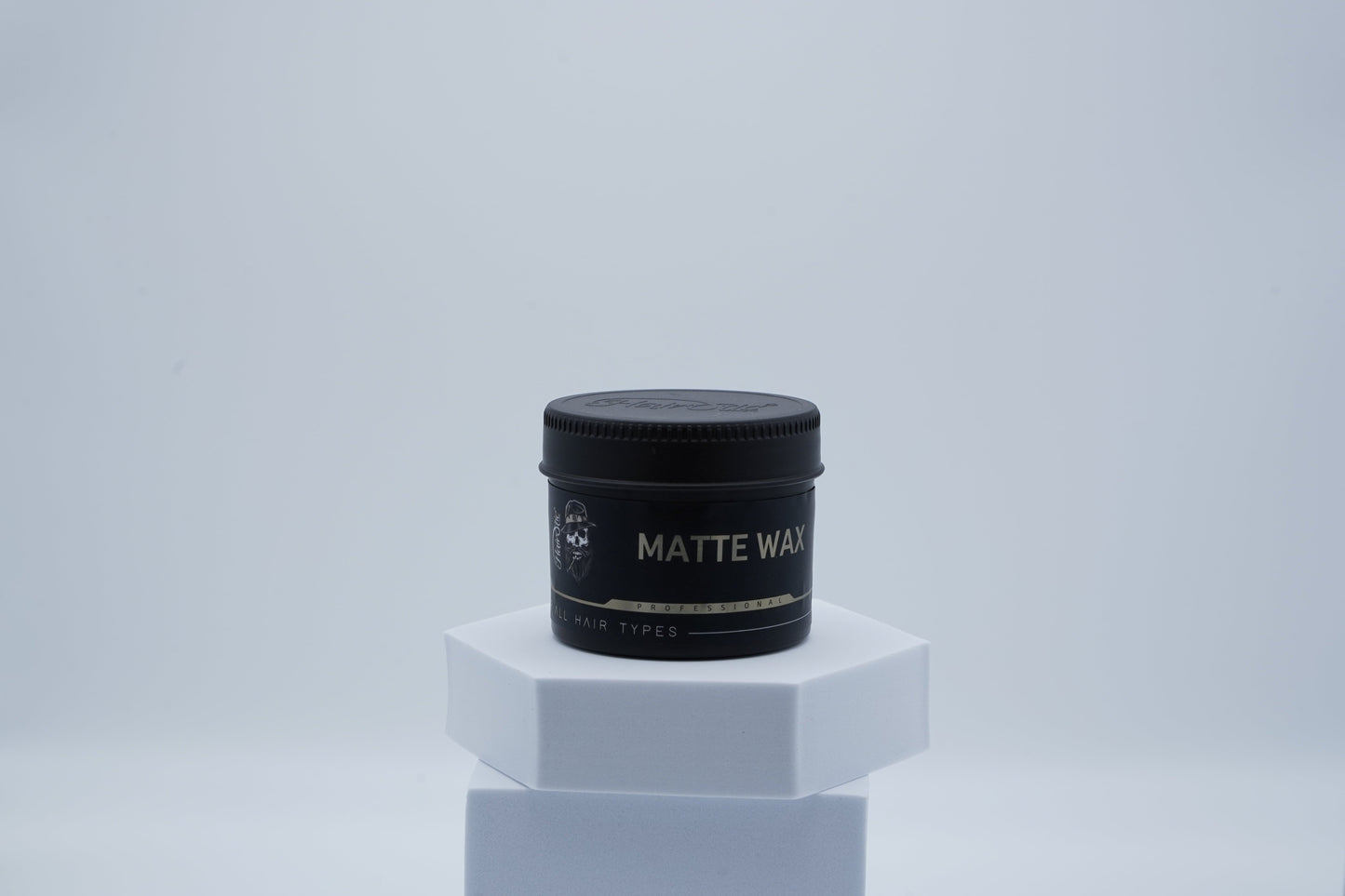 Schwarze Dose Hairotic Matte Wax mit goldenem Etikett für professionelles Haarstyling, geeignet für alle Haartypen, auf weißem Podest vor hellem Hintergrund.