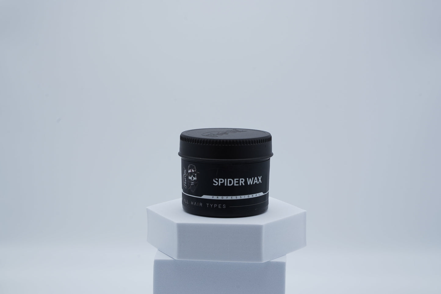Schwarze Dose Hairotic Spider Wax für alle Haartypen auf weißem Podest, vor hellem Hintergrund.