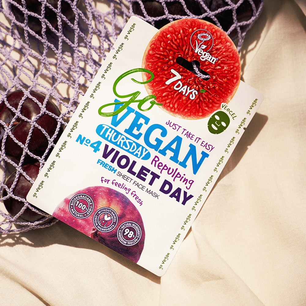 Packung der 7DAYS Go Vegan Thursday Violet Day Fresh Sheet Face Mask mit Feigen- und Pflaumenmotiv, veganem Label und natürlichem Netzbeutel im Hintergrund.