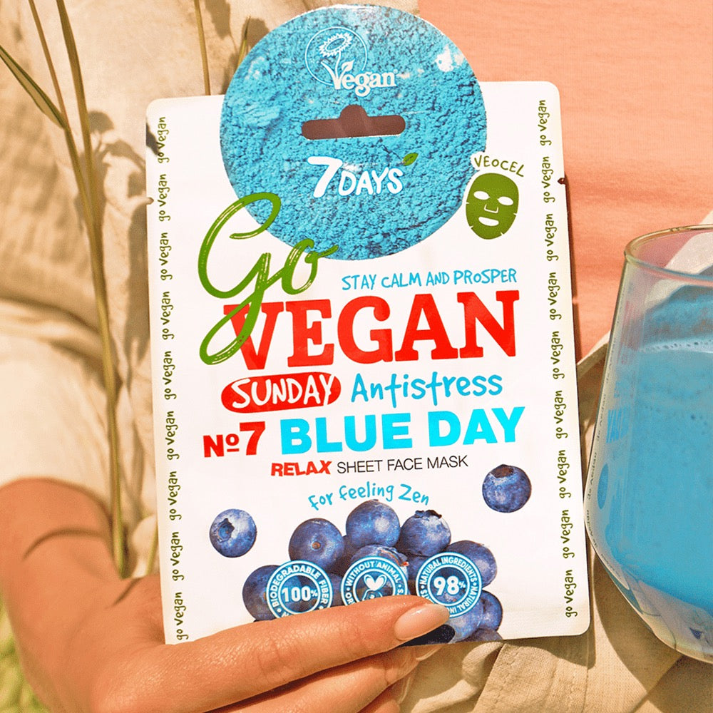 nd hält eine 7DAYS Go Vegan Sunday Antistress Sheet Face Mask Blue Day Packung mit Blaubeeren-Motiv, veganem Label und blauem Getränk daneben.
