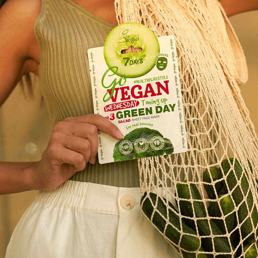Frau hält die 7DAYS Go Vegan Wednesday Green Day Salad Sheet Face Mask mit Gurken- und Salatmotiv in der Hand, daneben ein Netzbeutel mit frischen Gurken – veganes Label gut sichtbar.