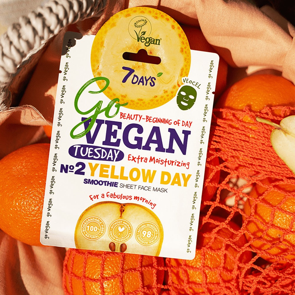 7DAYS Go Vegan Tuesday Yellow Day Smoothie Sheet Face Mask mit Maracuja- und Orangenmotiv, fotografiert in einem Netzbeutel voller Zitrusfrüchte – veganes Label deutlich sichtbar.
