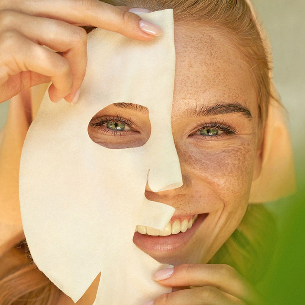 Junge Frau mit Sommersprossen lächelt in die Kamera, während sie sich eine weiße Sheet-Maske vorsichtig auf das Gesicht auflegt.