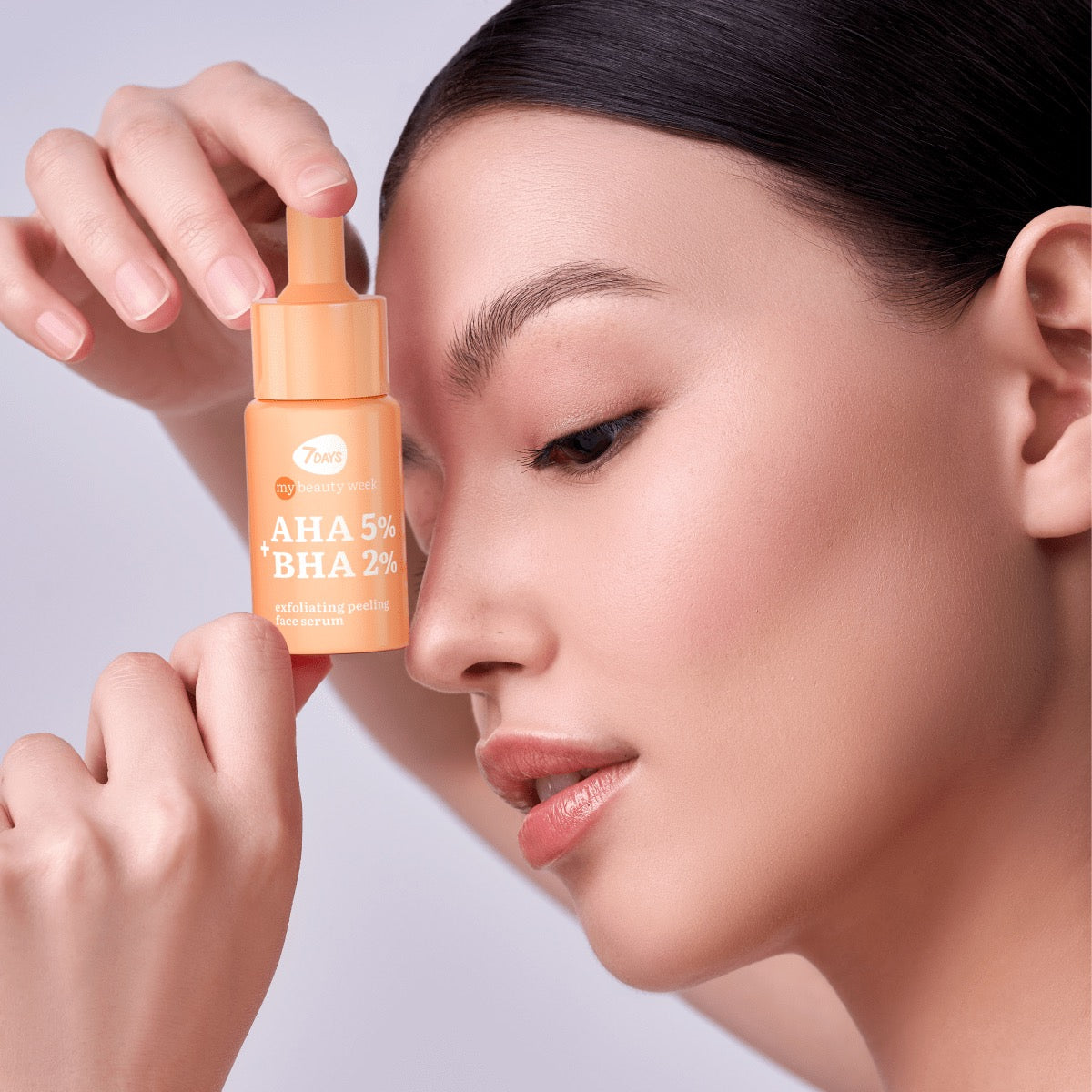 Frau mit glatter Haut und natürlichem Make-up hält ein oranges Fläschchen 7DAYS AHA 5% + BHA 2% Exfoliating Peeling Face Serum nah an ihr Gesicht.