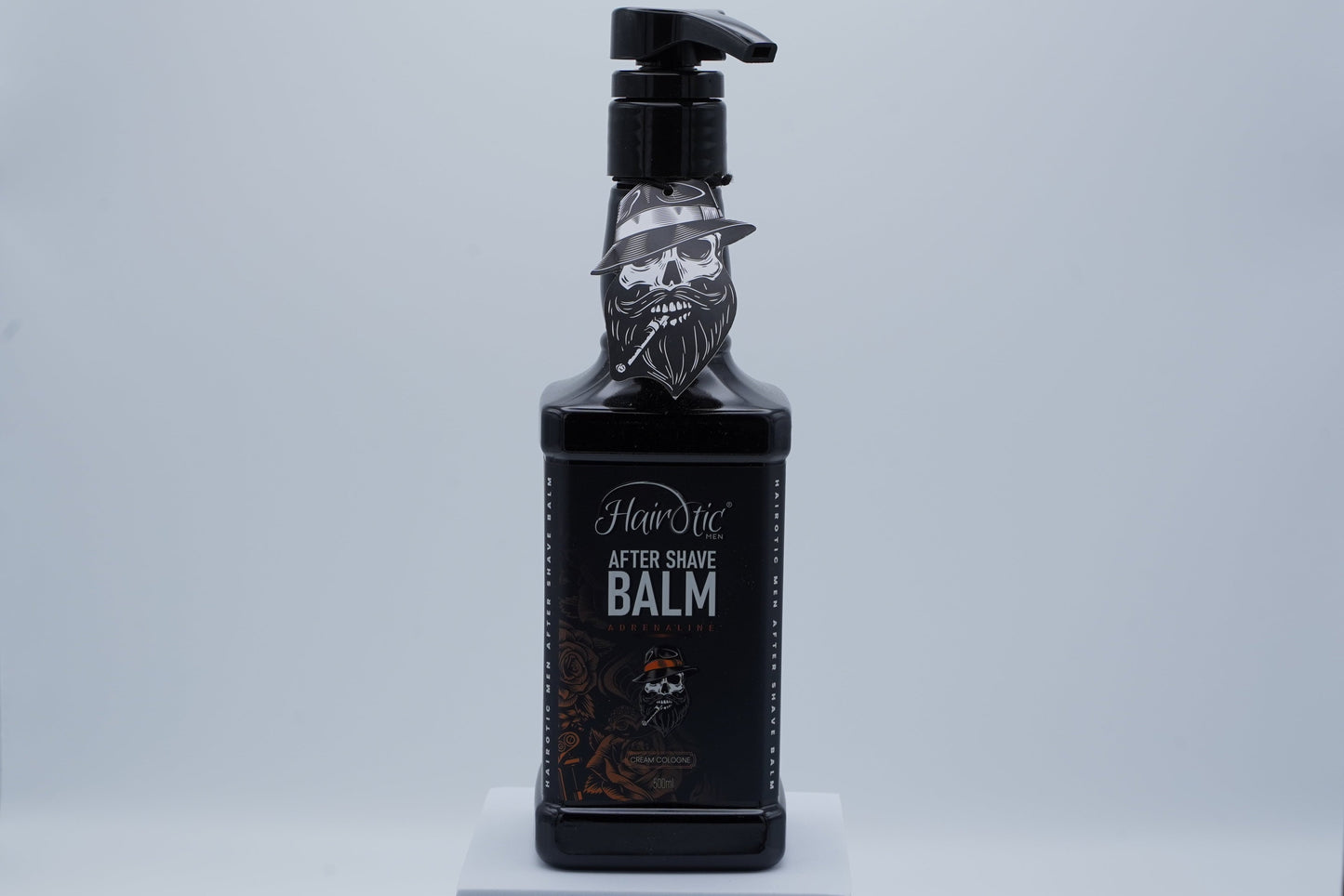 Schwarze Pumpflasche Hairotic After Shave Balm Adrenaline, mit Totenkopf-Logo, stehend auf weißem Podest vor neutralem Hintergrund.