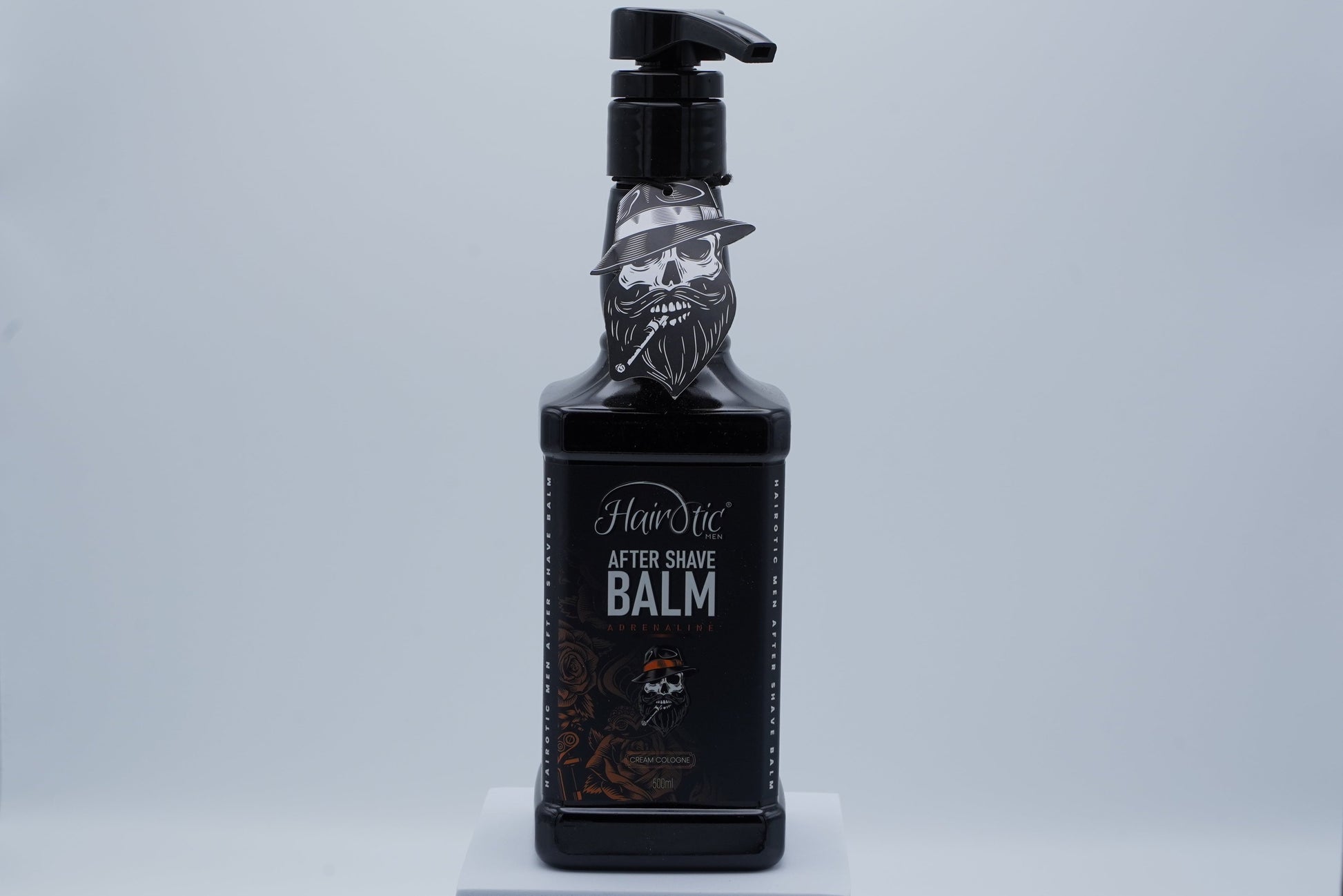 Schwarze Pumpflasche Hairotic After Shave Balm Adrenaline, mit Totenkopf-Logo, stehend auf weißem Podest vor neutralem Hintergrund.