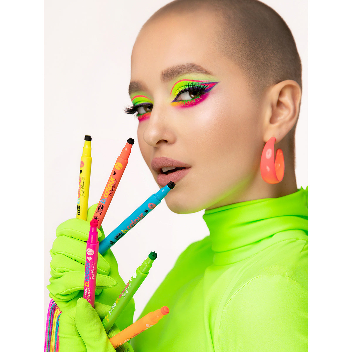 Model mit auffälligem Neon-Make-up und grünem Outfit präsentiert fünf b.colour Eyeliner & Stamp Pens in verschiedenen Neonfarben, ideal für kreative Eyeliner-Looks mit Stempelmotiven.